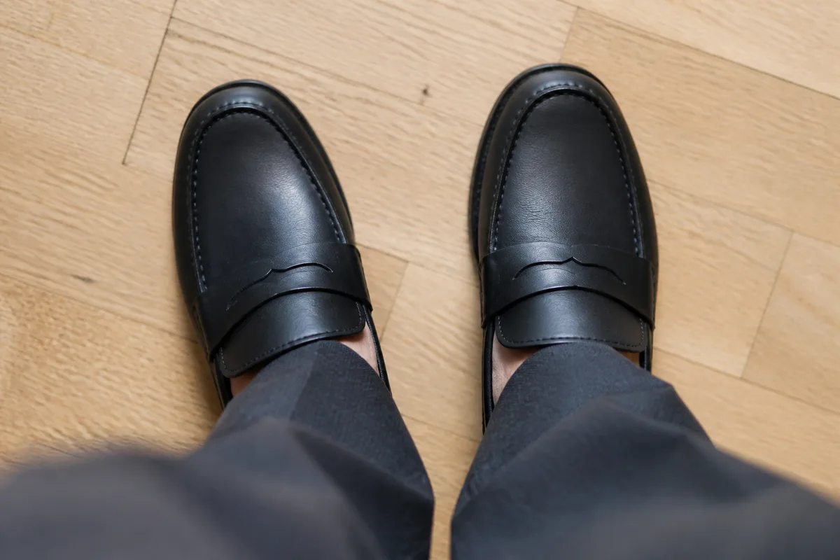 Paire de mocassins en cuir noir posée sur un parquet clair à côté d&rsquo;un bas de pantalon à pli